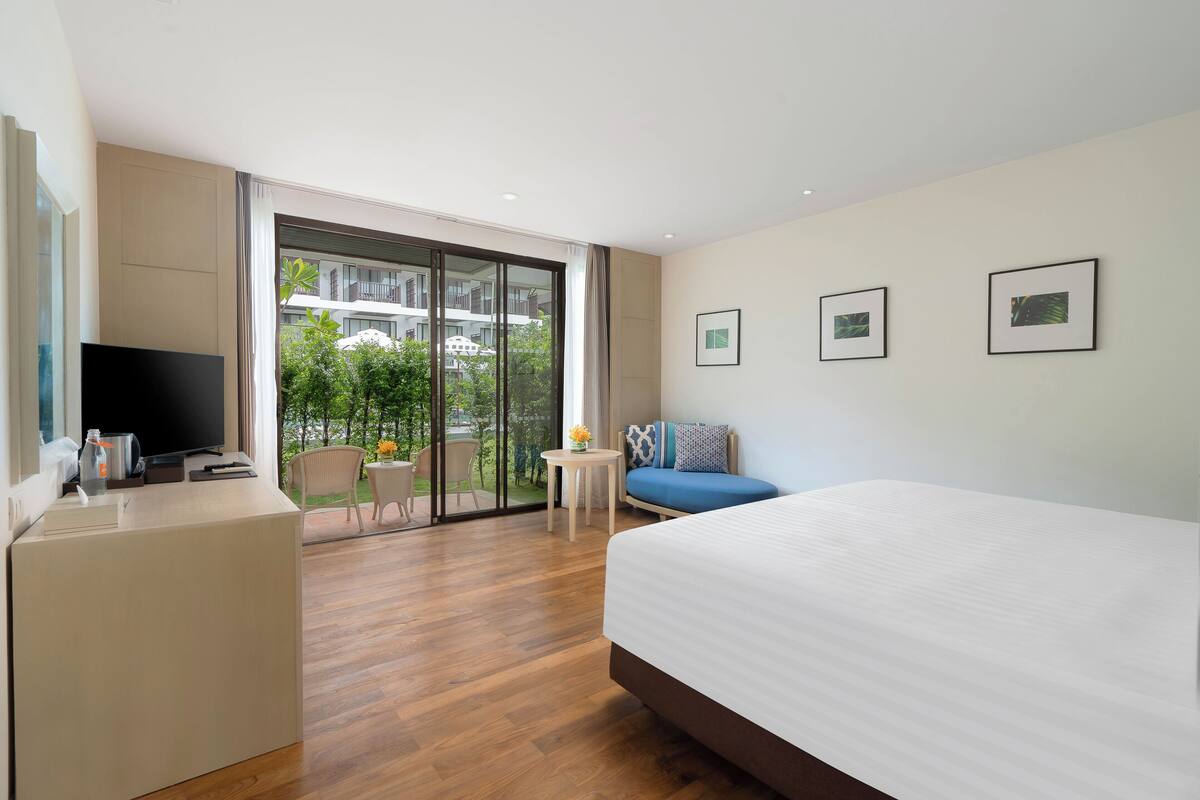 Et moderne hotelværelse med en stor seng, et skrivebord med et fladskærms-tv, et lille bord med en vase, og en balkon med udsigt til grønne områder og bygninger.