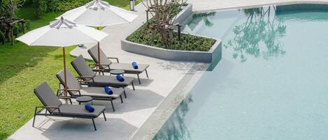 A poolside area with lounge chairs and umbrellas, surrounded by greenery and a clear pool.