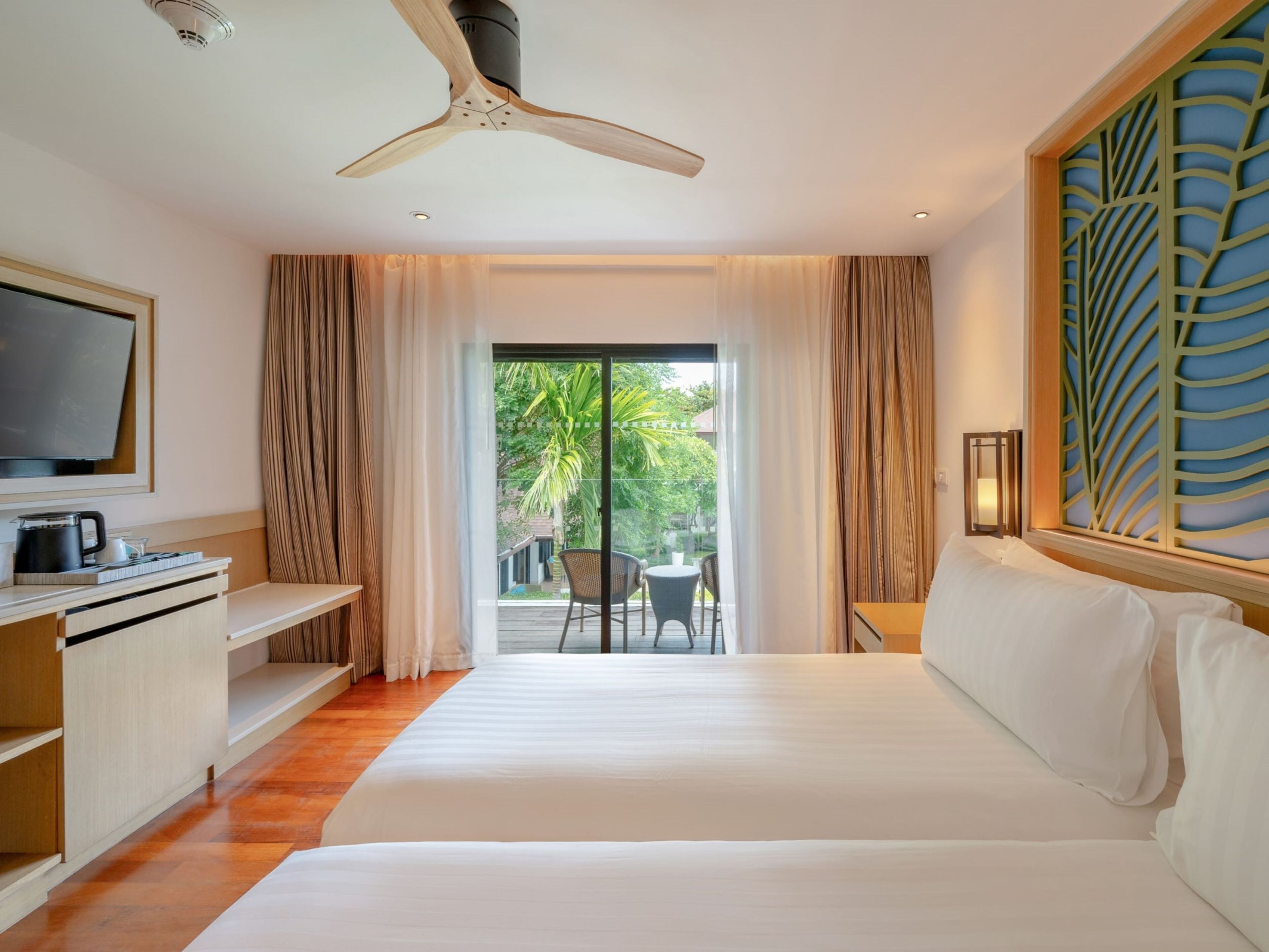 A hotel room with a bed, a desk with a television, a ceiling fan, and a view of the outdoors.