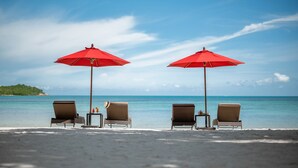 Deux chaises de plage, placées sous des parasols rouges, offrent une vue sur l’océan et une île lointaine.