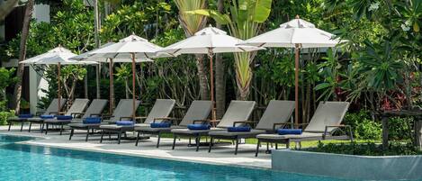 Zona junto a la piscina con sillones de descanso y sombrillas, rodeada de exuberante vegetación.