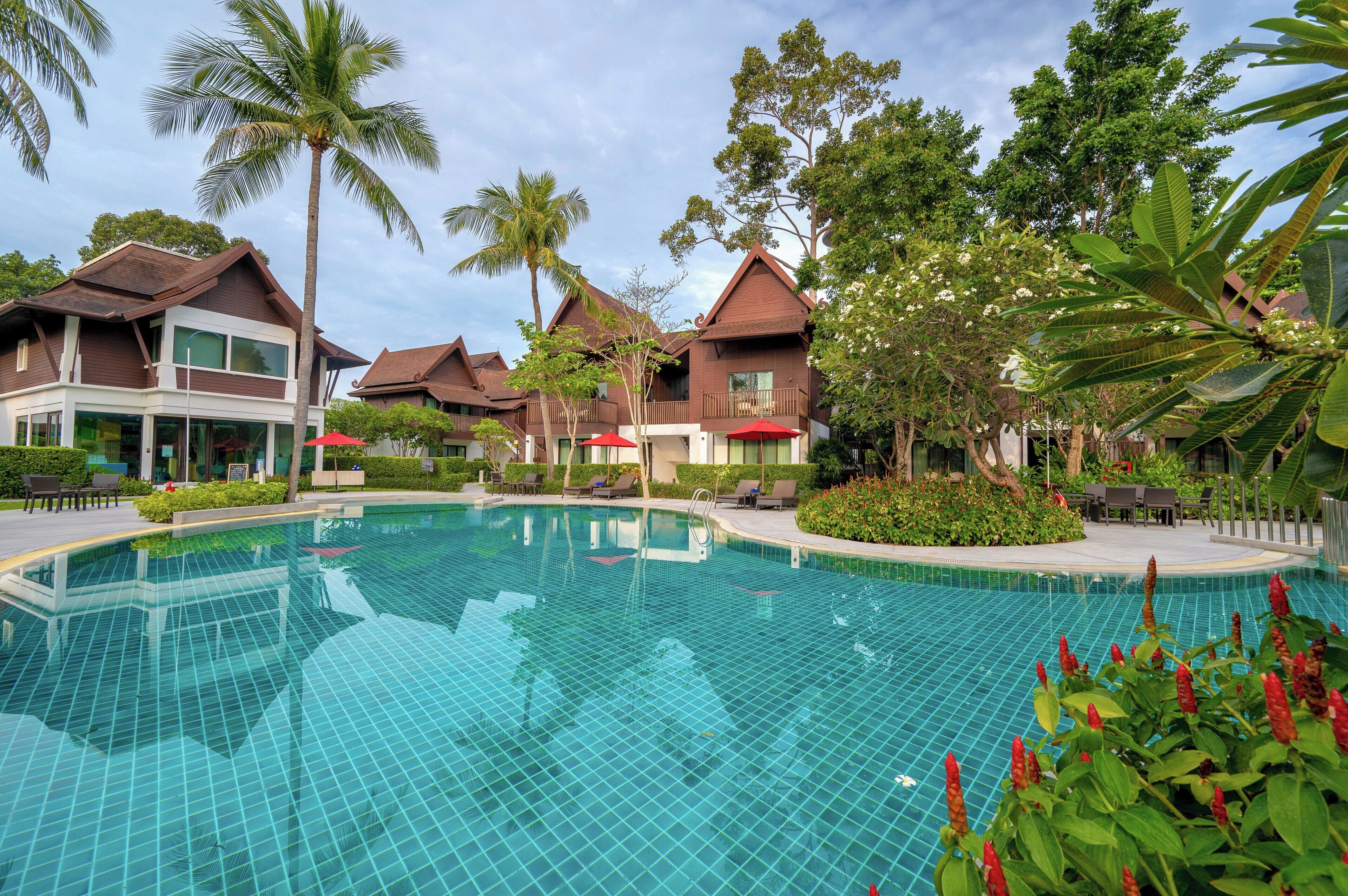 A swimming pool with tiled flooring, surrounded by lounge chairs and umbrellas, with buildings and palm trees in the background.