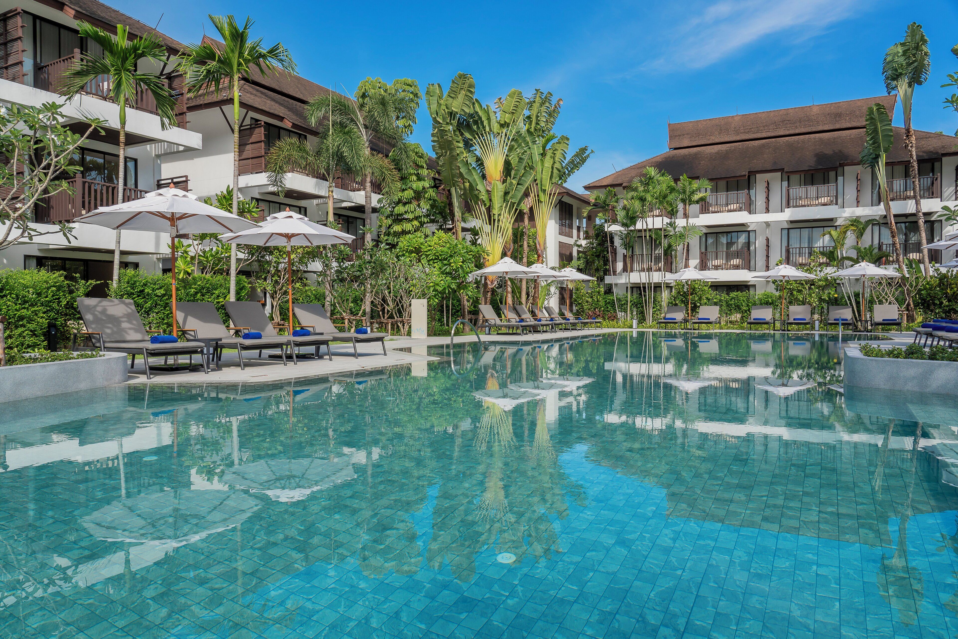 Un espace piscine d’hôtel avec des chaises longues et des parasols, entouré de palmiers et surplombé d’un ciel bleu azur.