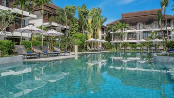 Un espace piscine d’hôtel avec des chaises longues et des parasols, entouré de palmiers et surplombé d’un ciel bleu azur.