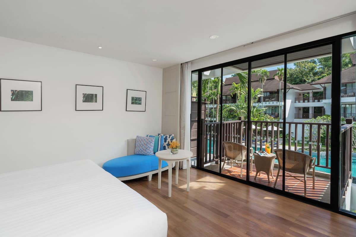 Et moderne hotelværelse med en stor seng, en blå sofa, et lille rundt bord med en vase, samt en balkon med pool og udendørs møbler.