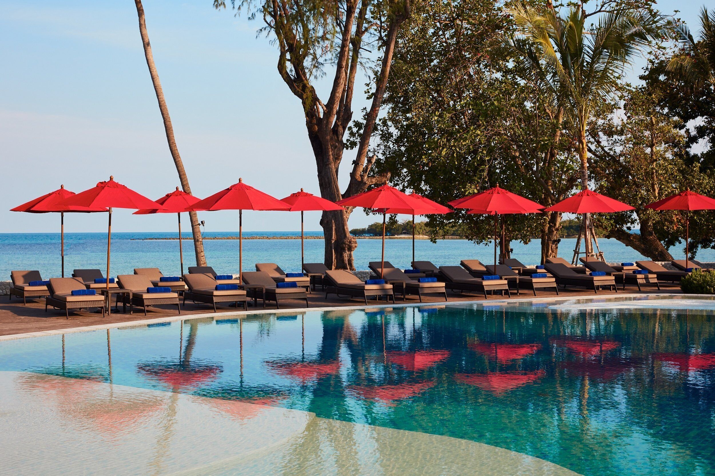 Zona de piscina con sillas de descanso y sombrillas rojas, rodeada de palmeras y vista al mar.