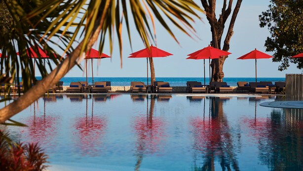 Un espace piscine agrémenté de chaises longues et de parasols rouges, qui se fond dans le paysage grâce aux palmiers environnants et offre une vue dégagée sur l’océan.
