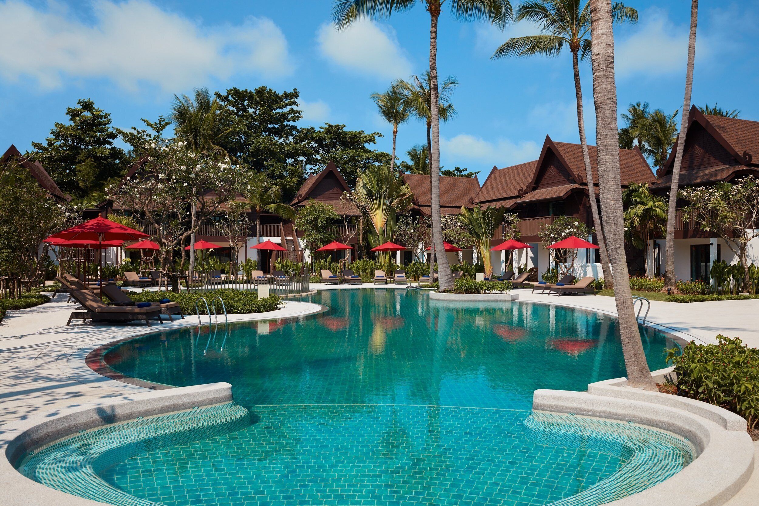 A resort pool with lounge chairs and umbrellas, surrounded by palm trees and traditional architecture.