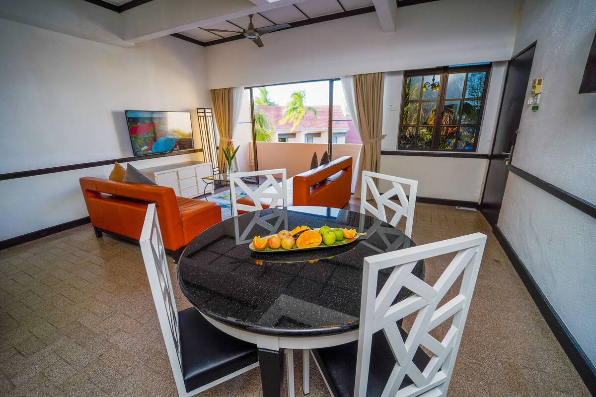 A dining area with a round table, white chairs, and a fruit platter.