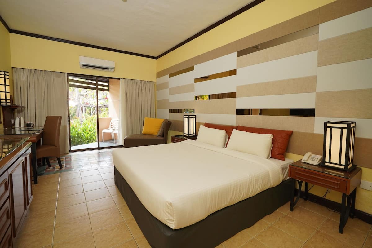 A hotel room with a bed, a desk, a chair, a nightstand with a lamp, and an air conditioning unit.