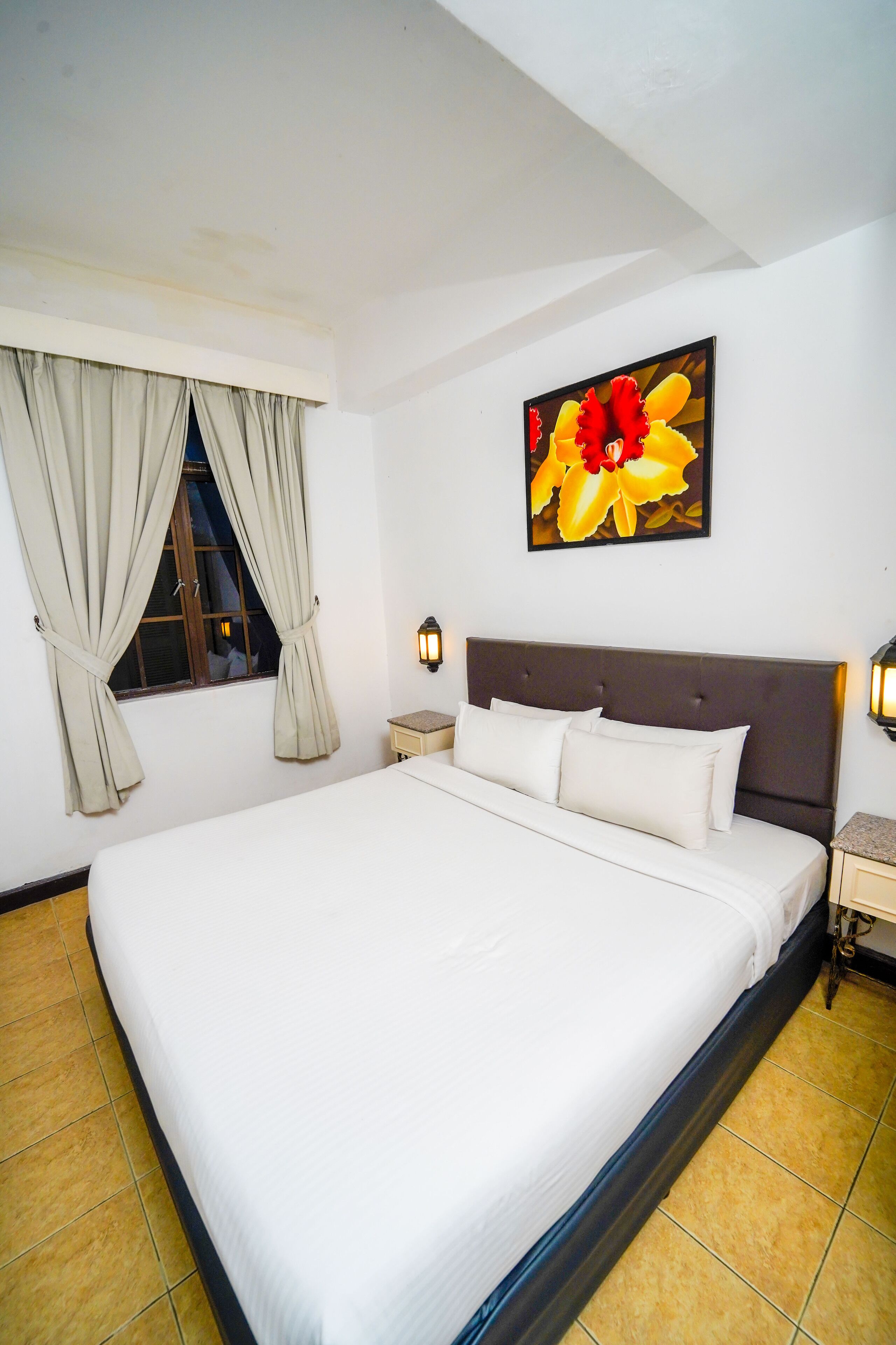 A neatly made bed with a headboard, a window with curtains, a wall-mounted light, and a painting of flowers.