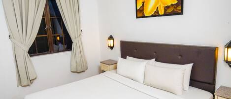 A neatly made bed with a headboard, a window with curtains, a wall-mounted light, and a painting of flowers.