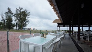 An outdoor dining area with white tables and chairs, a thatched roof, and a view of the beach and ocean. - Holiday Villa Beach Resort & Spa Cherating (Cherating)