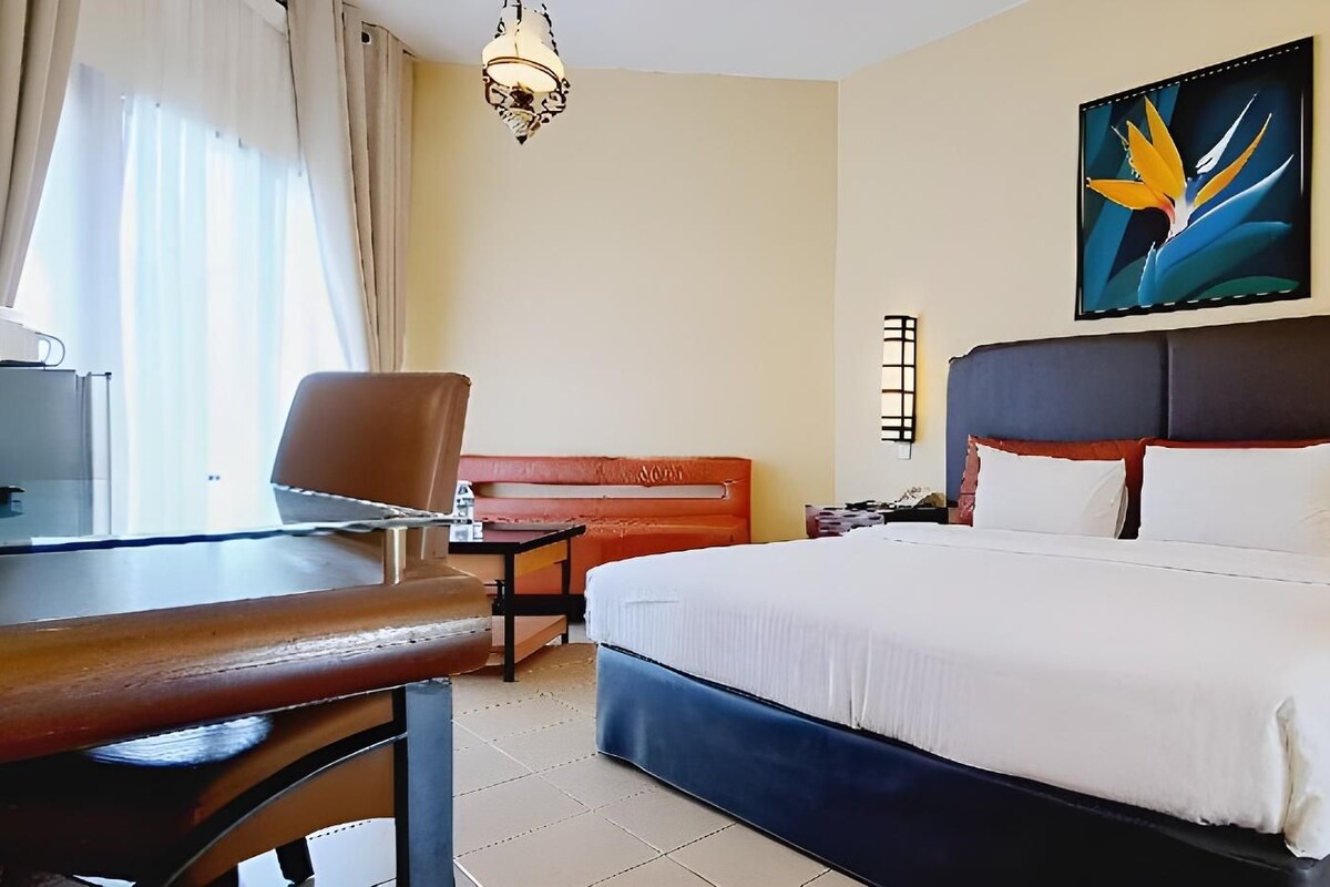A hotel room with a bed, a desk, a chair, a sofa, and a painting on the wall.