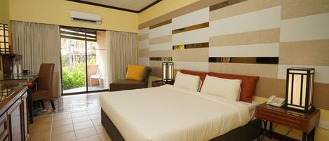 A hotel room with a large bed, a desk with a chair, a small table, and a nightstand with a lamp.