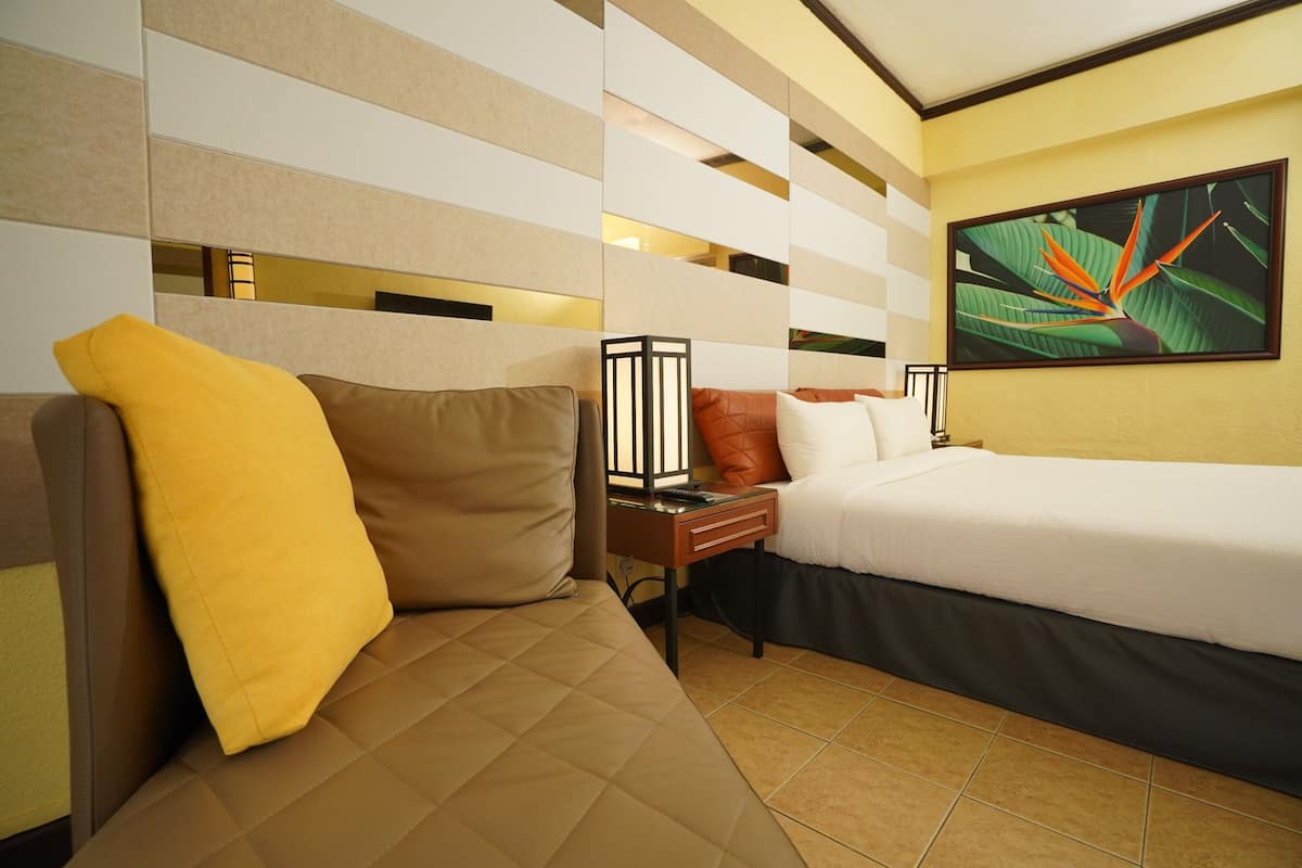 A hotel room with a bed, a sofa, a nightstand, a lamp, and a painting on the wall.