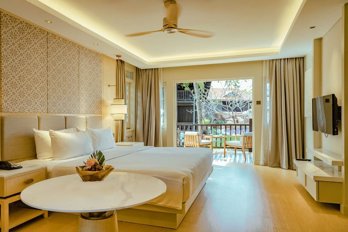 Quarto de hotel com uma cama grande, mesa de cabeceira com abajur, uma pequena mesa com uma cesta de frutas, televisão e varanda com cadeiras.