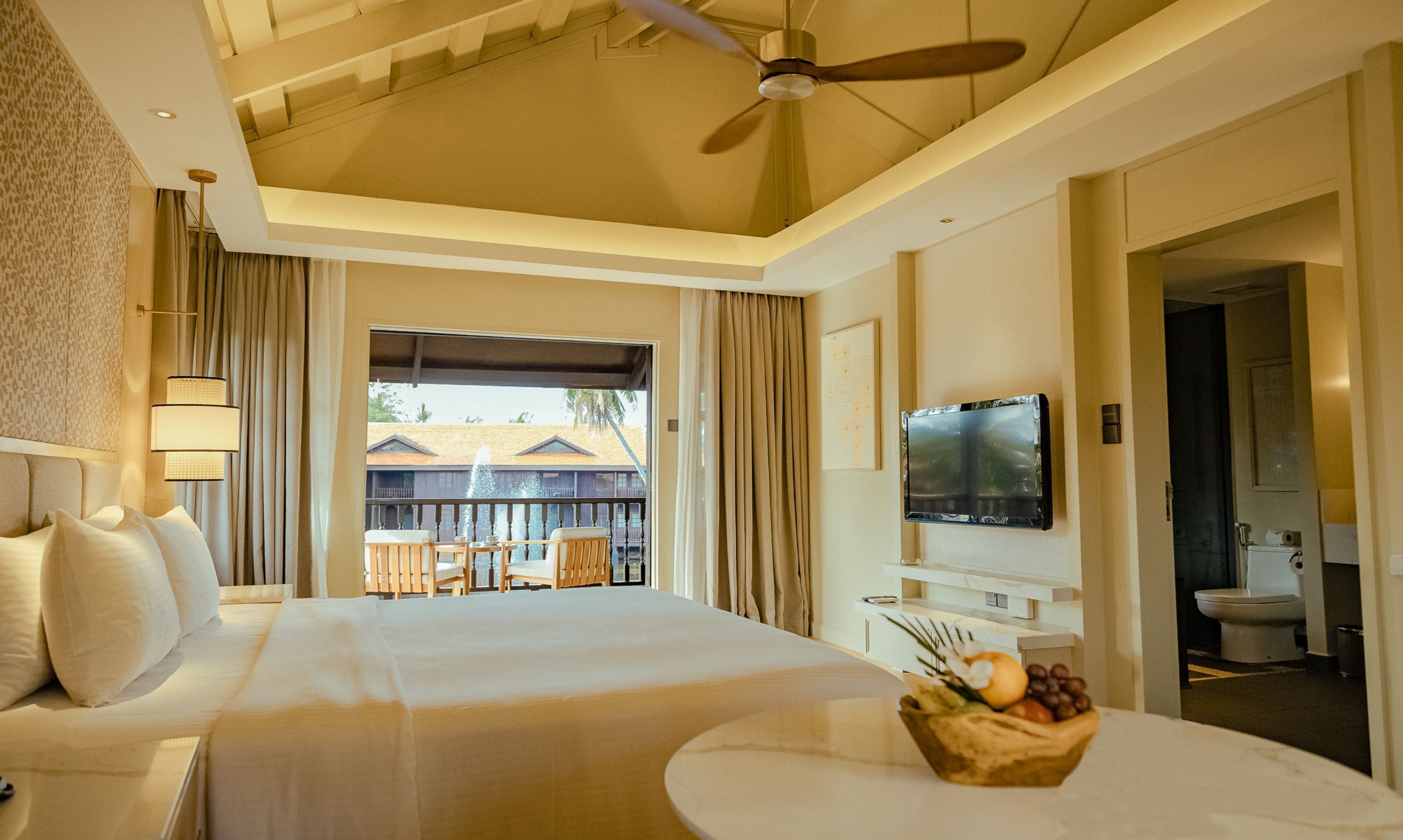 A hotel room with a large bed, a ceiling fan, a TV, and a balcony with a view of buildings.