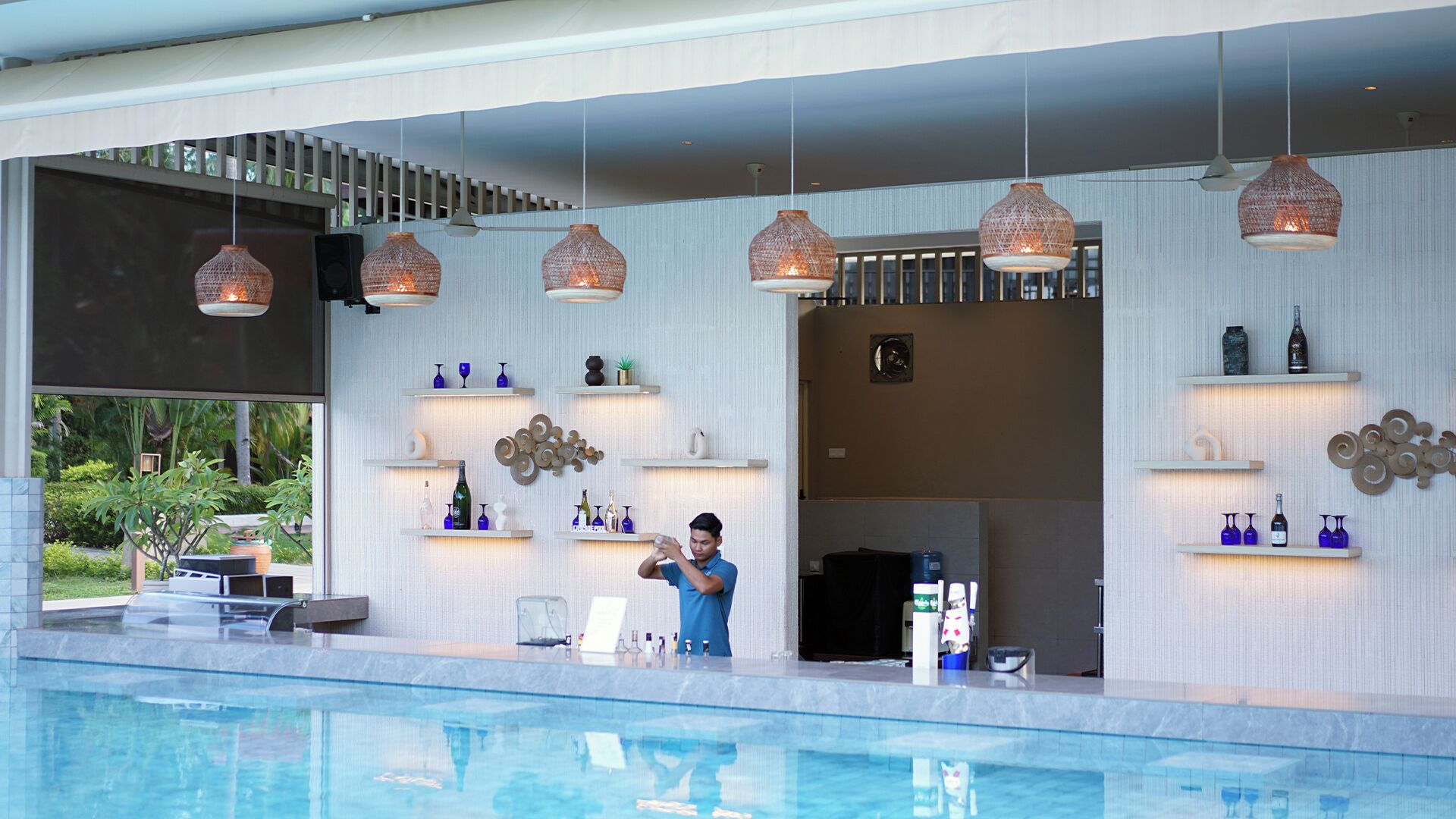 Een poolbar met hangende lampen, flessen op planken, en een persoon die bij het zwembad staat.