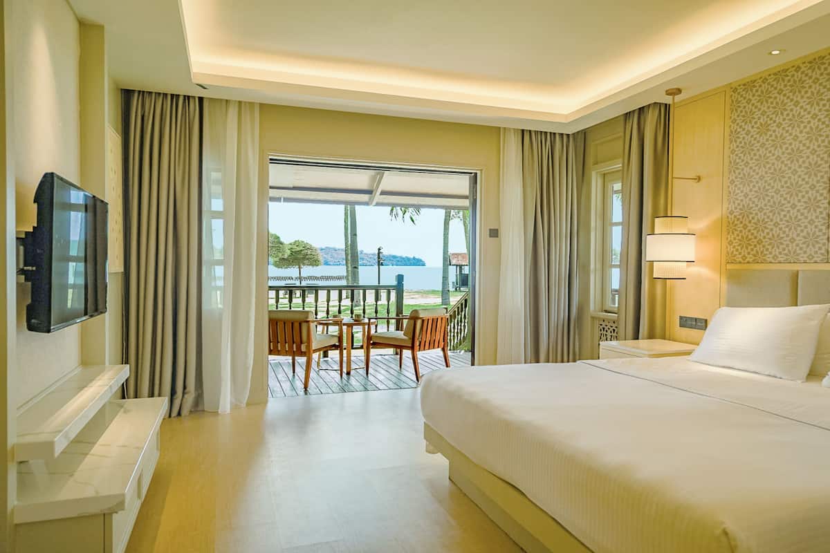 Quarto de hotel com uma cama grande, televisão, varanda com mesa e cadeiras, e vista para o mar.