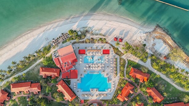 Vista aérea de un complejo turístico con una piscina central, varias edificaciones de techo rojo y una zona de playa.