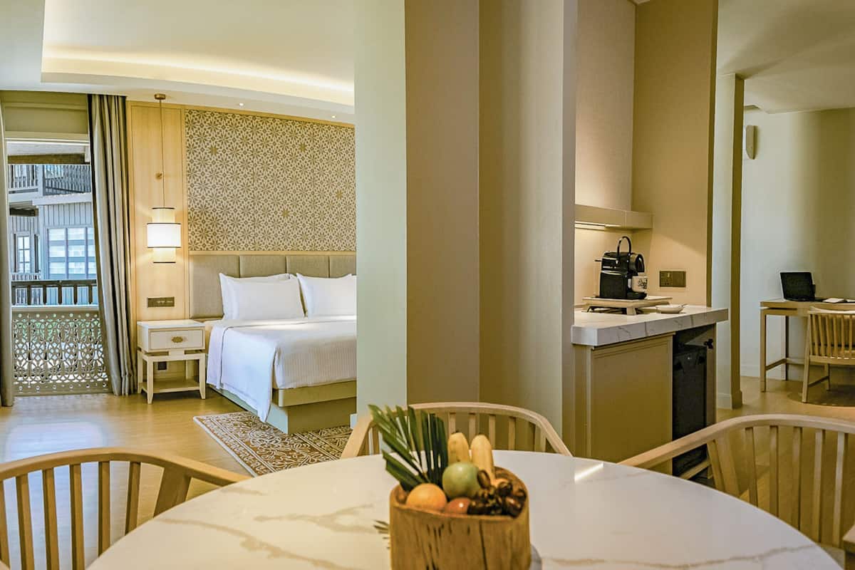 Quarto de hotel com cama, escrivaninha, cadeira e mesa de jantar com frutas.