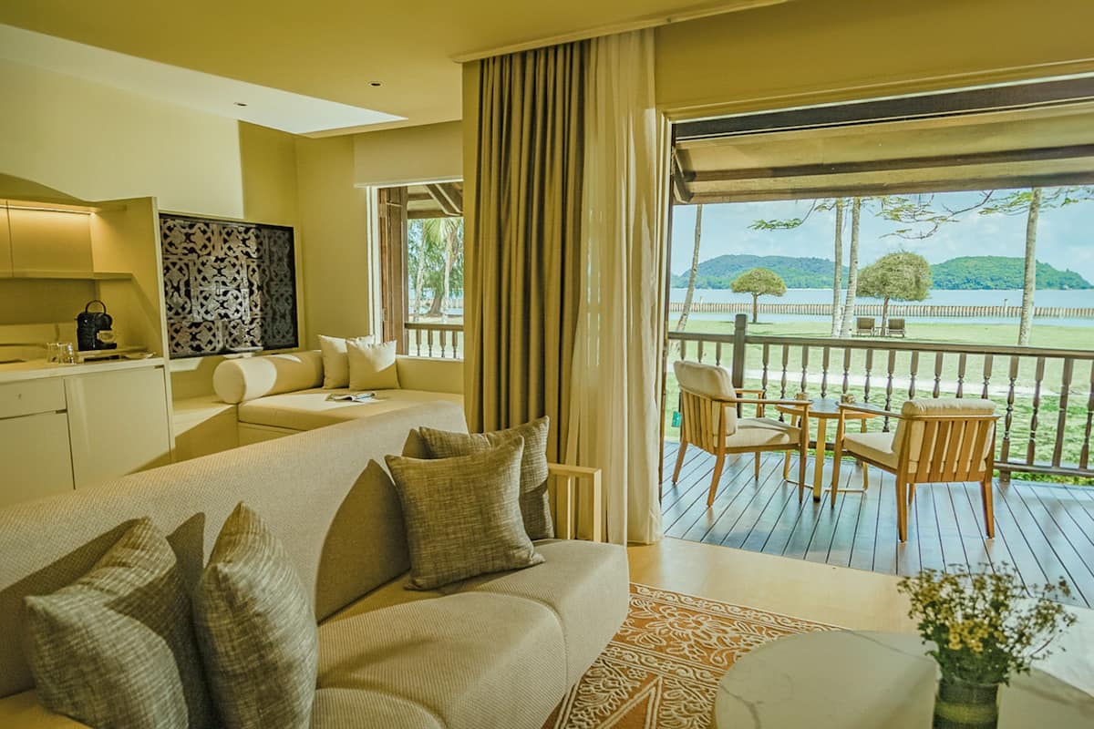 Quarto de hotel moderno com um sofá grande, área de jantar com cadeiras, cozinha e varanda com vista para o mar.