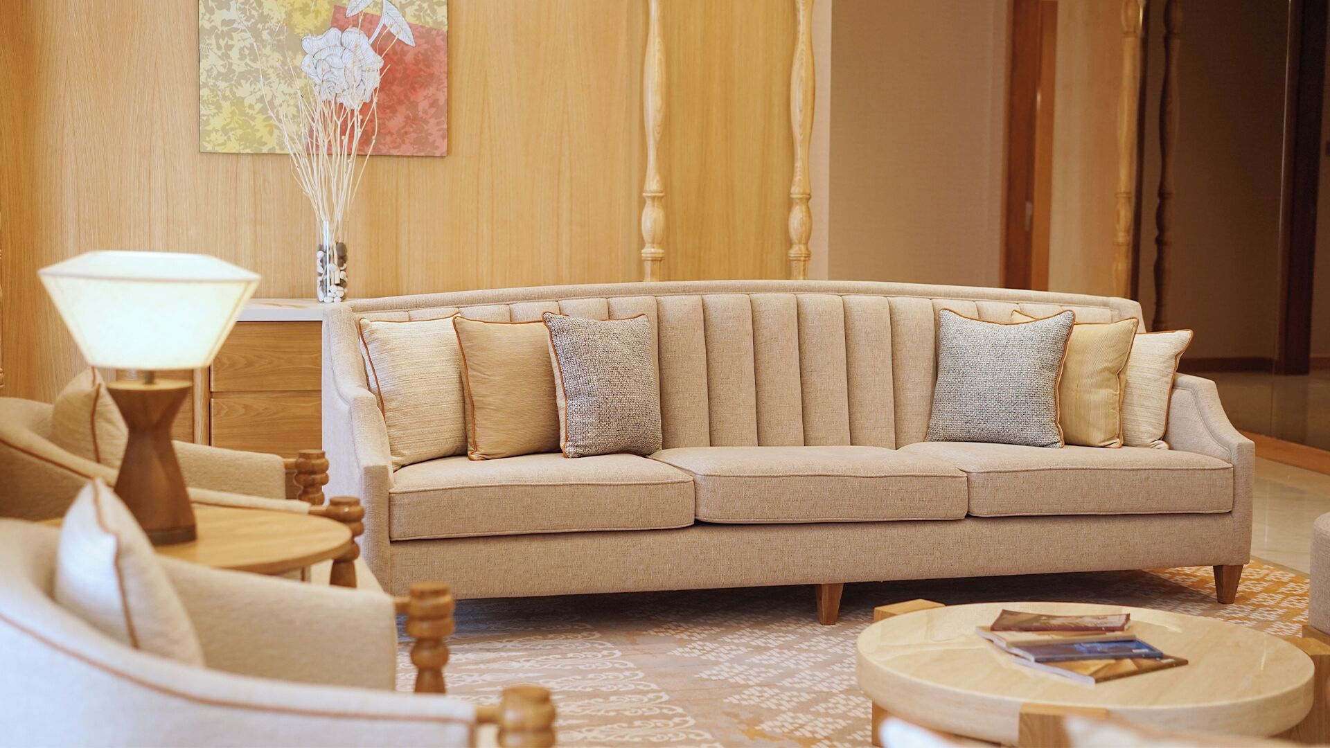 A beige sofa with two pillows, a round wooden coffee table, a floor lamp, and a patterned rug in a well-lit room with wooden paneling.
