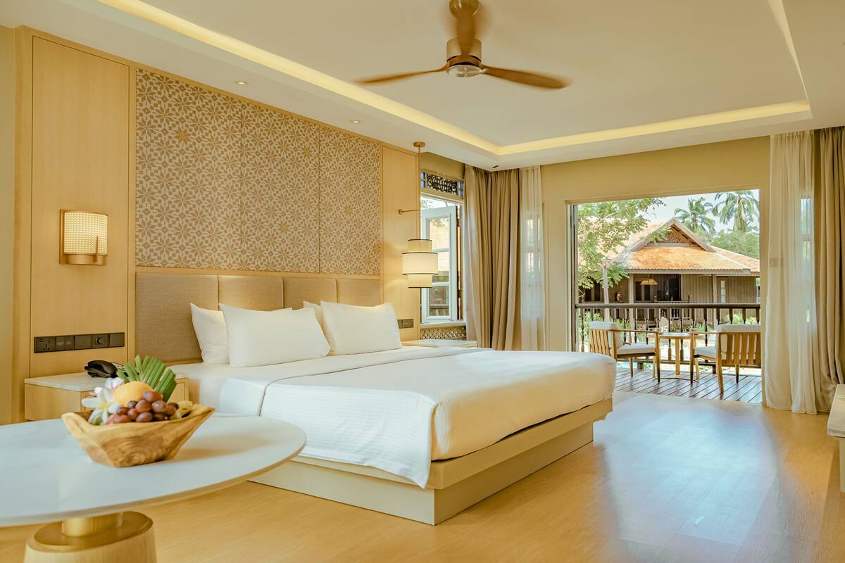 Quarto de hotel com uma cama grande, uma mesa, uma cadeira e uma varanda com vista para uma casa.