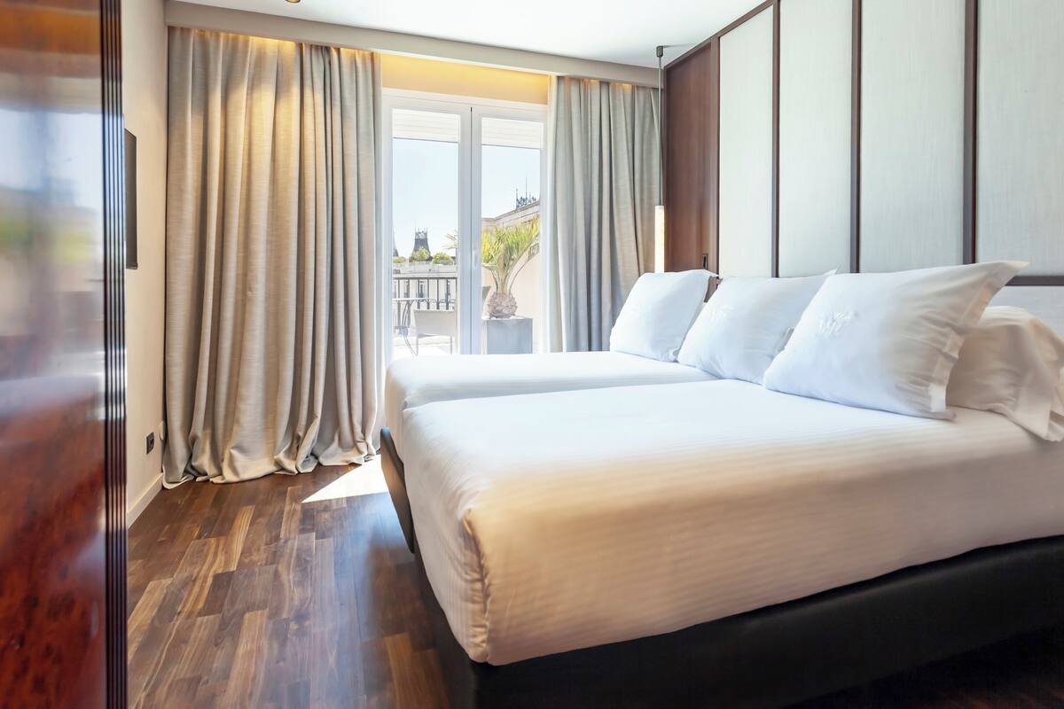 A hotel room with a large bed, a sliding glass door leading to a balcony, and a wooden floor.