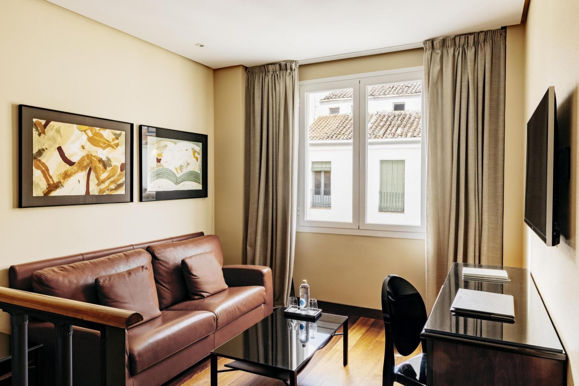 A modern hotel room with a leather sofa, a glass coffee table, a desk with a chair, two framed artworks, a large window with curtains, and a flat-screen TV mounted on the wall.