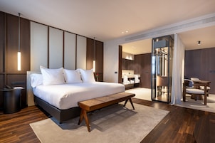 A modern hotel room with a large bed, bedside table, and a bench. There is a bathroom with a mirror and a shower area.