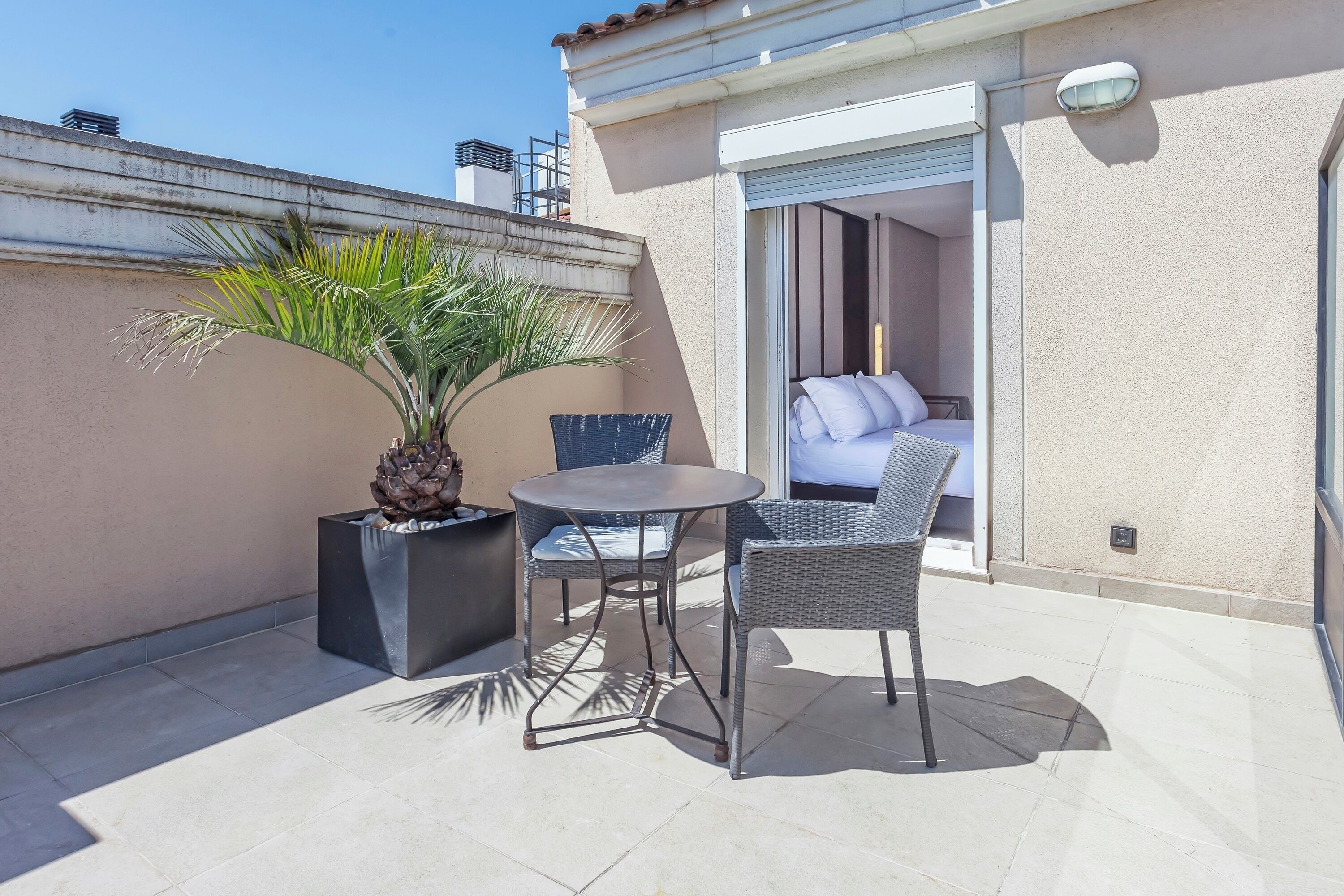 A small patio with a round table and two wicker chairs, a potted plant, and a sliding glass door leading to a bedroom with a bed and pillows.
