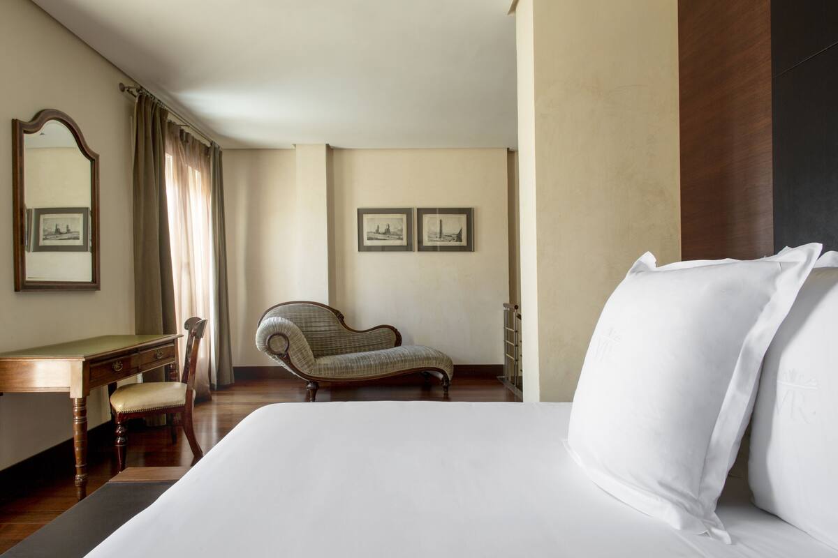 A hotel room with a large bed, a wooden desk, a chair, and two framed pictures on the wall.