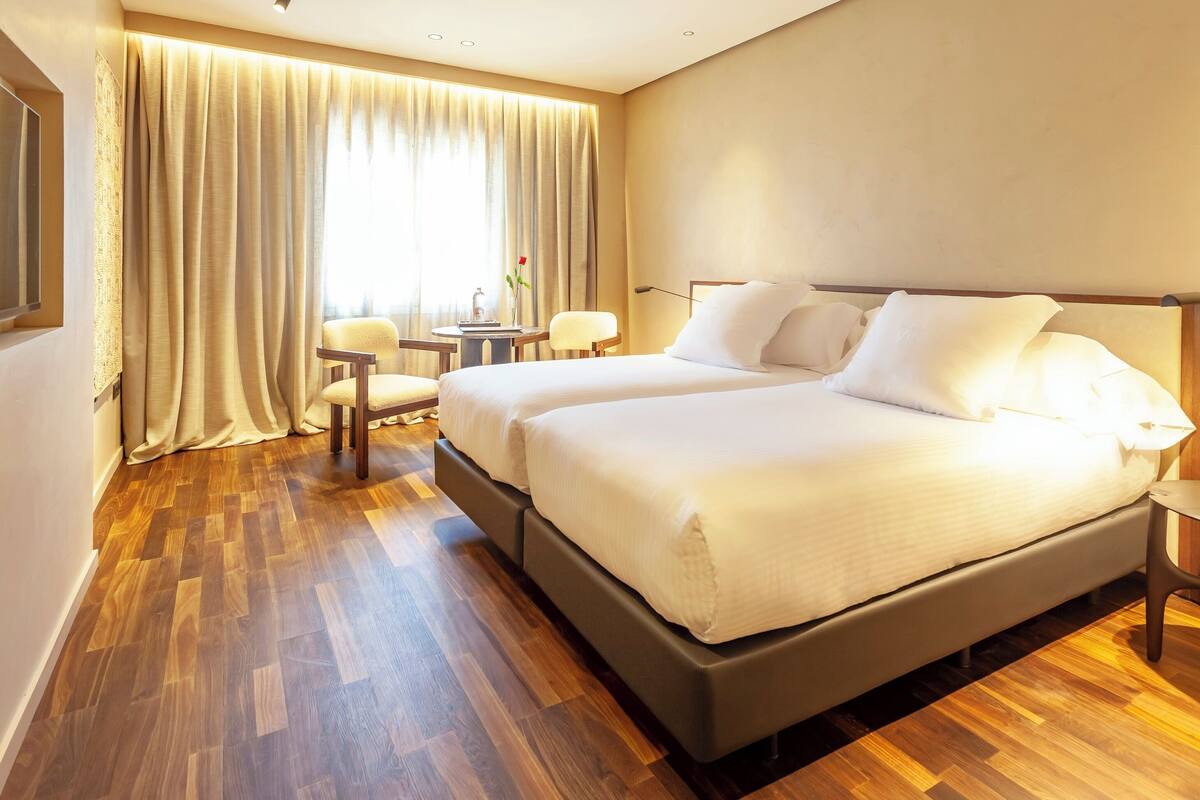 A hotel room with a large bed, two bedside tables, a chair, a small table, and a window with curtains.