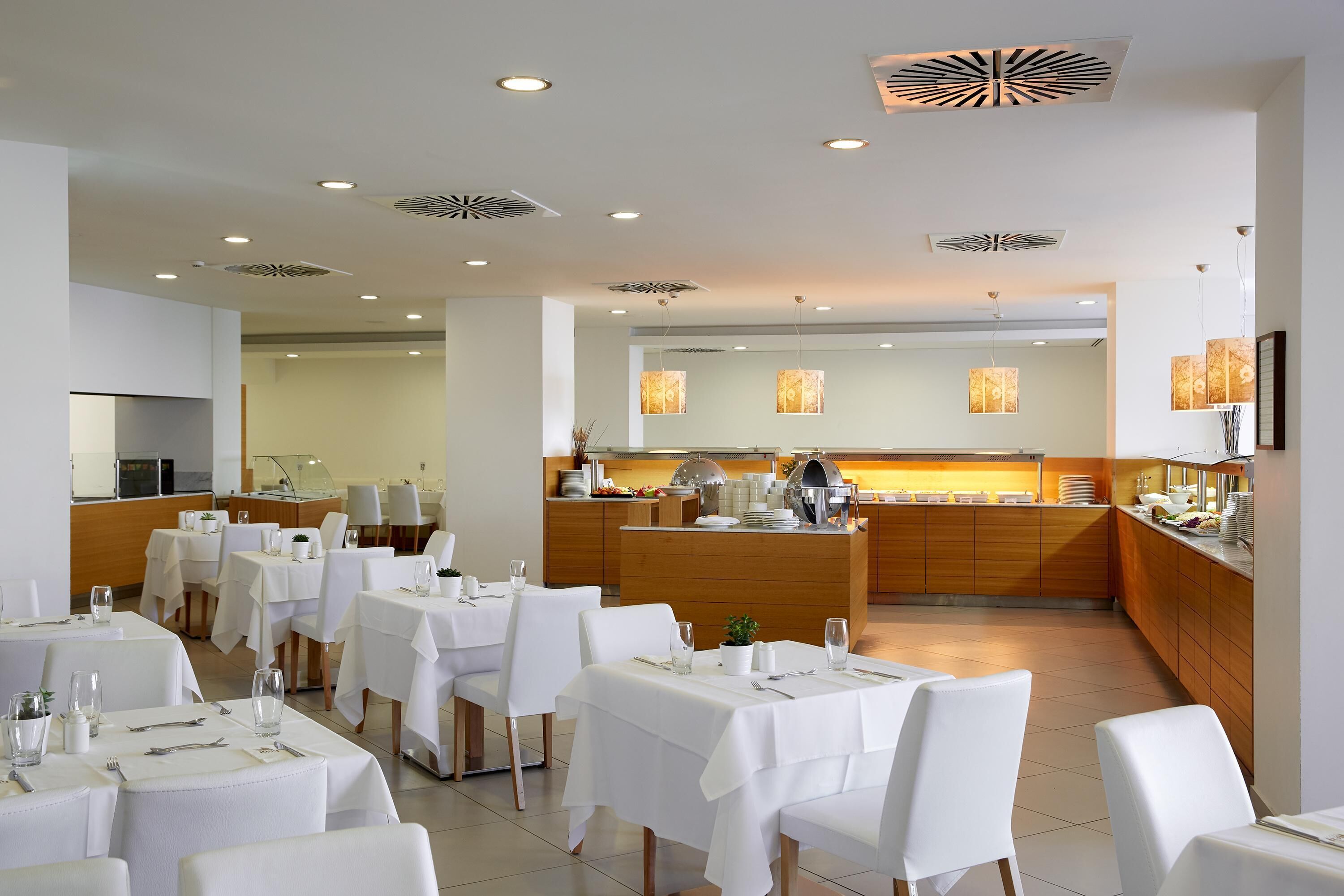 Un espace repas moderne avec des tables et des chaises blanches, un îlot central servant de buffet et des spots encastrés au plafond.