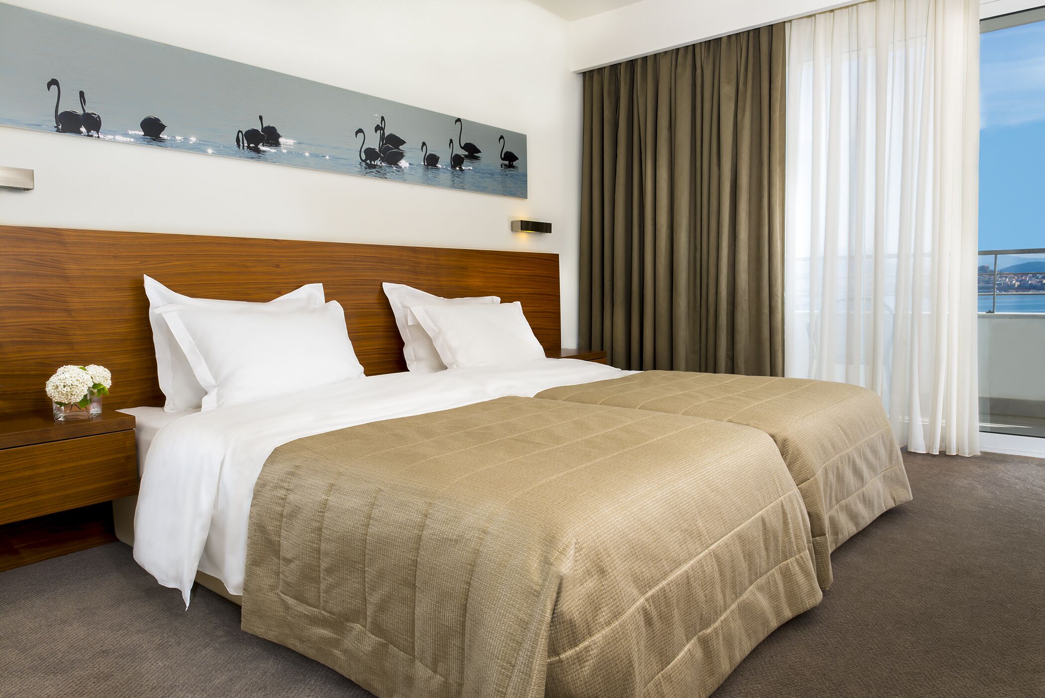 A hotel room with a large bed, a wooden headboard, a painting of swans, a nightstand with a vase of flowers, and a view of the sea through the window.