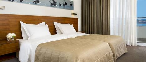 A hotel room with a large bed, a wooden headboard, a painting of swans, a nightstand with a vase of flowers, and a view of the sea through the window.