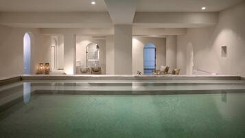A large indoor swimming pool with a tiled floor, surrounded by seating areas and a glass partition.