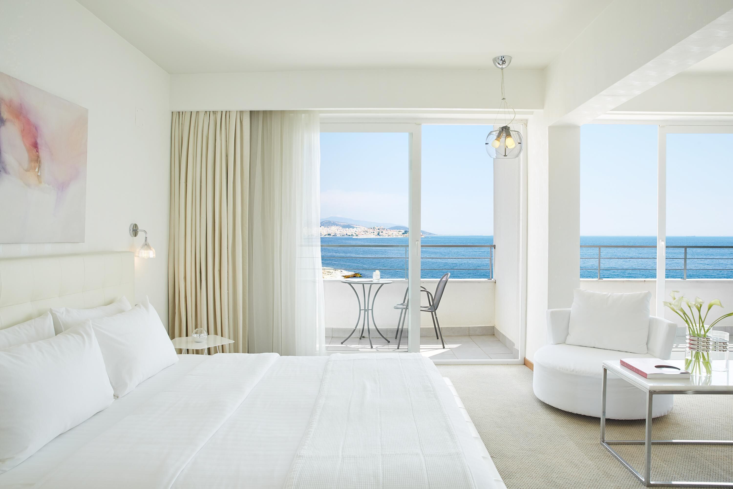 Ein Hotelzimmer mit einem großen Bett, einem Balkon mit Meerblick, einem kleinen Tisch und einem Stuhl.