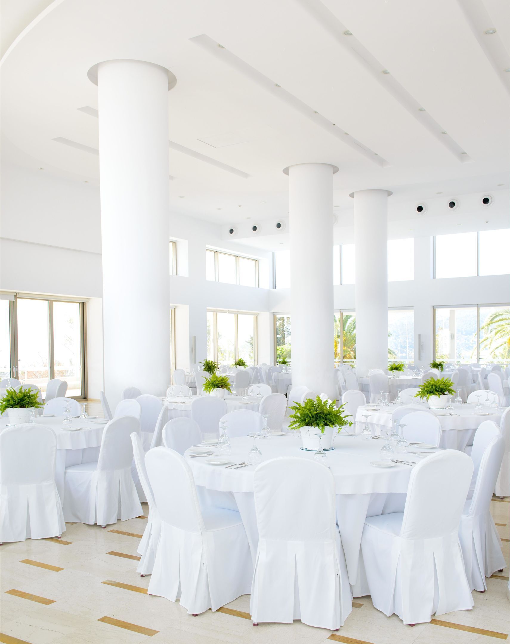 Une salle de banquet spacieuse, avec des tables et des chaises blanches, de grandes fenêtres et des colonnes blanches.