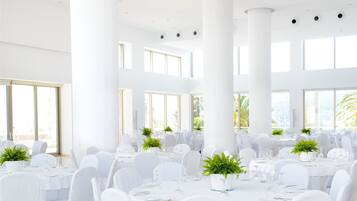 Un amplio salón de banquetes con mesas y sillas blancas, ventanas grandes y columnas blancas.