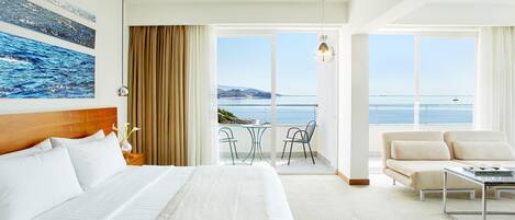 A hotel room with a large bed, a sofa, a balcony with a view of the sea, and a dining area.