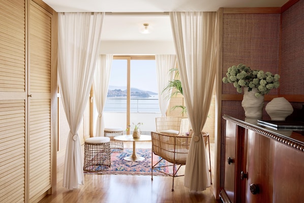 Una habitación con piso de madera, una mesa redonda con sillas, vista al mar y un mueble con objetos decorativos.