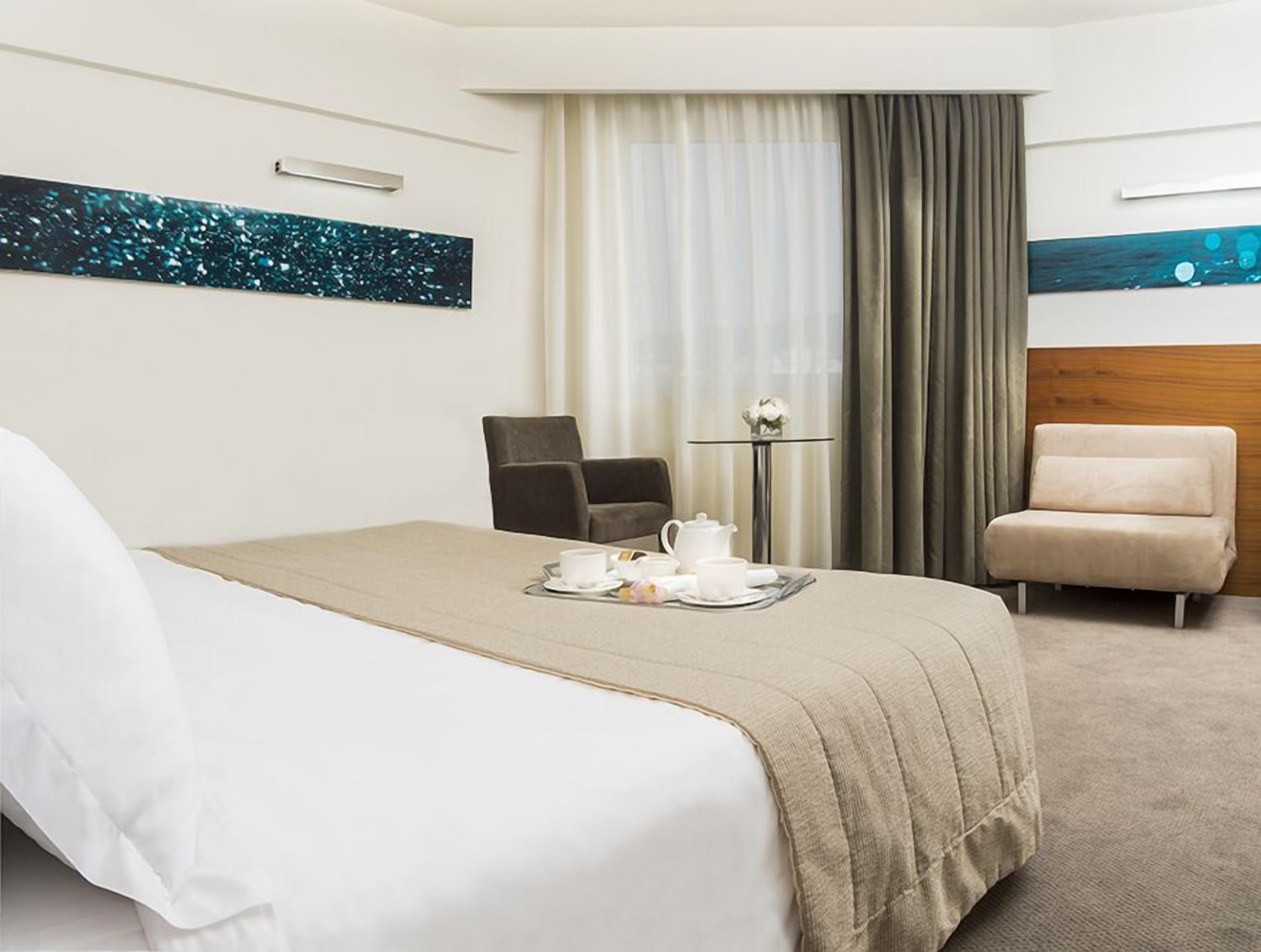 A hotel room with a bed, a chair, a small table with a vase, and artwork on the walls.