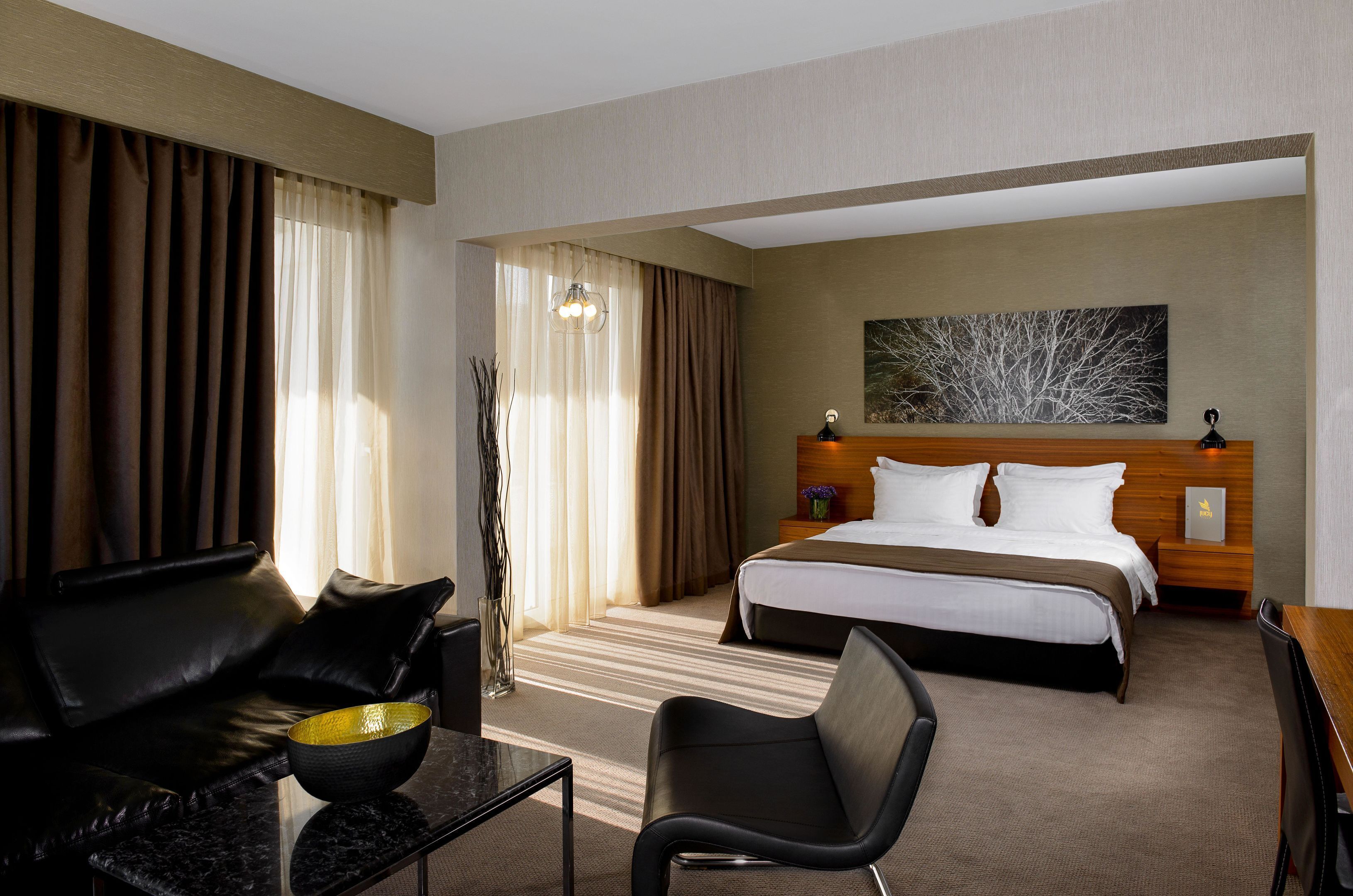 A hotel room with a bed, a desk, a chair, a coffee table, and a large window with curtains.