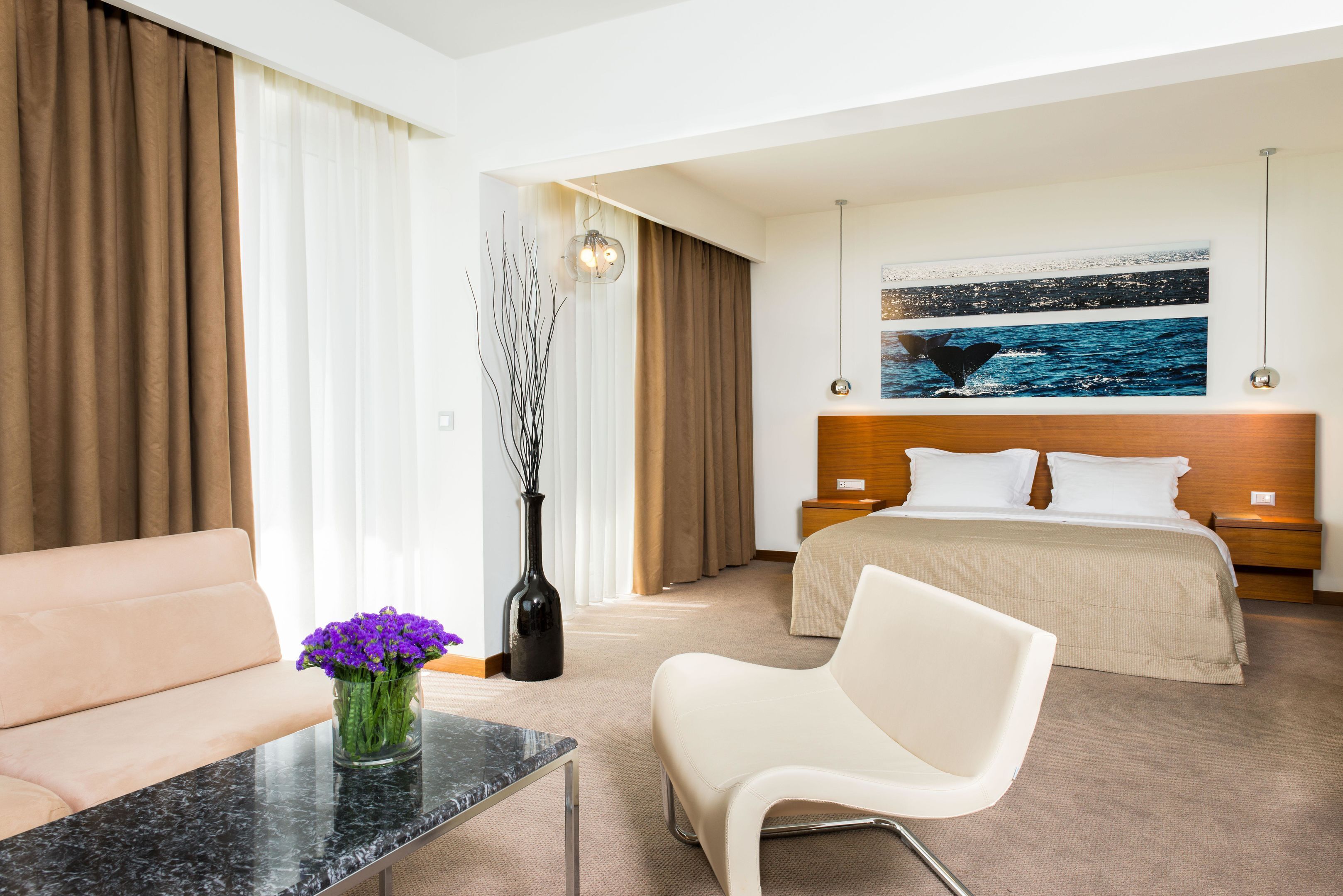 A hotel room with a bed, a sofa, a chair, a coffee table, and a vase with flowers.