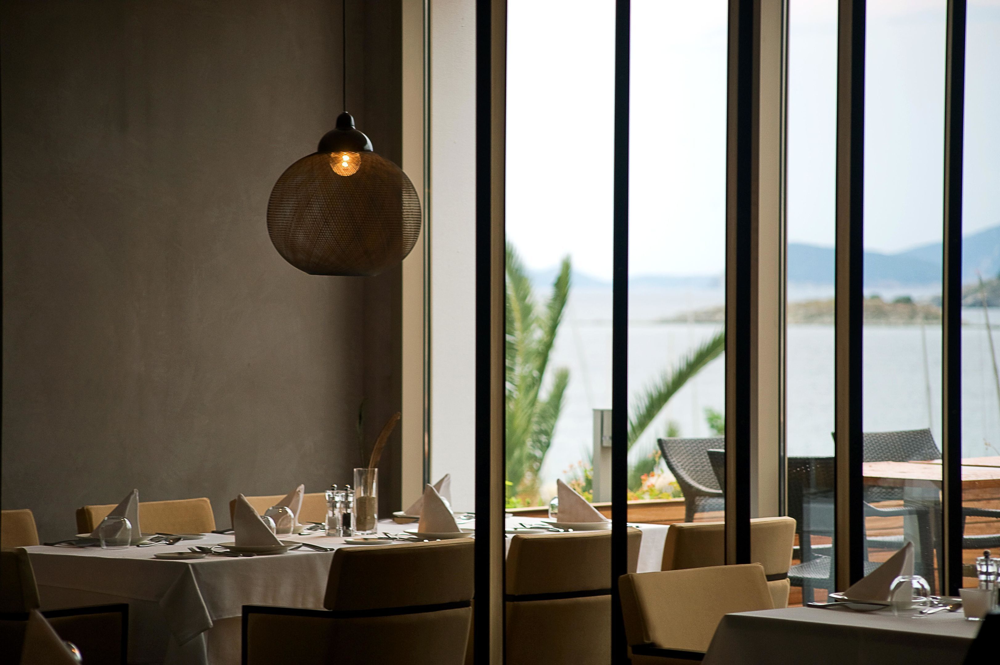 Un restaurant avec des tables dressées pour le service, de grandes fenêtres offrant une vue sur la mer et les montagnes, et un luminaire suspendu.