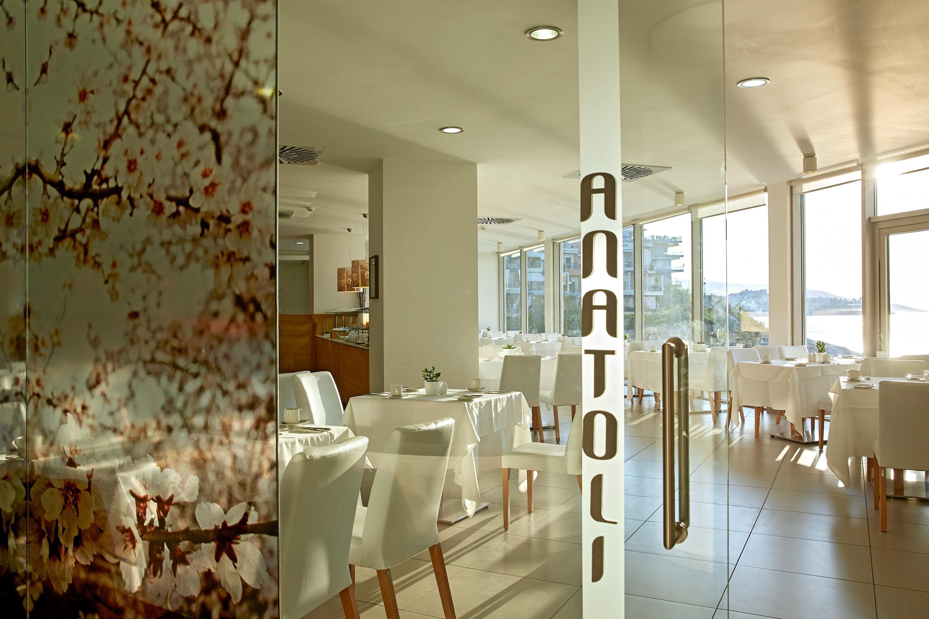 Una zona d'esmorzar moderna amb taules i cadires blanques, grans finestres que ofereixen una vista de la ciutat i una paret decorativa amb motius florals.