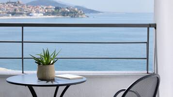 A balcony with a round table, a potted plant, and a chair. A view of the sea and a distant town is visible through the railing.